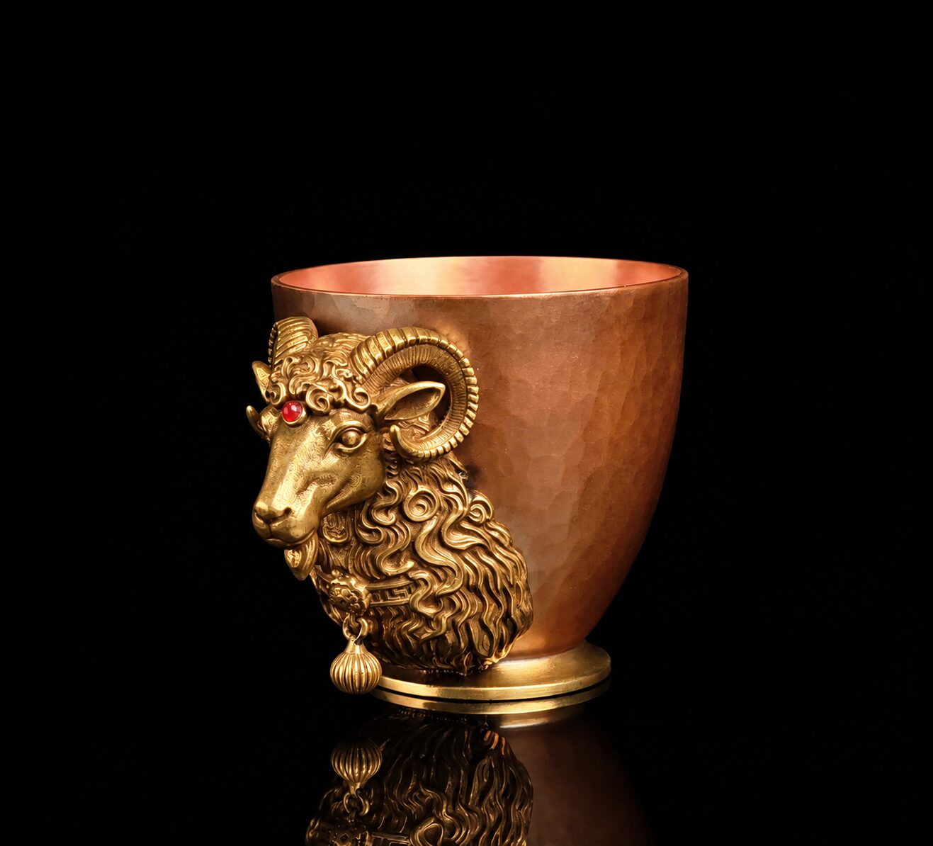 Three-quarter view of hammered red-copper mini cup with brass ram-head relief on black background