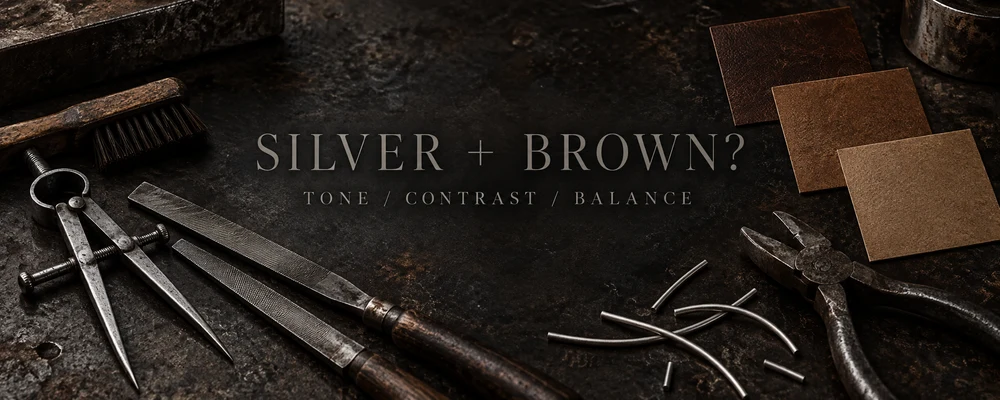 Silver and brown outfit contrast represented by steel tools, raw silver wire and warm brown swatch cards on a dark workshop bench