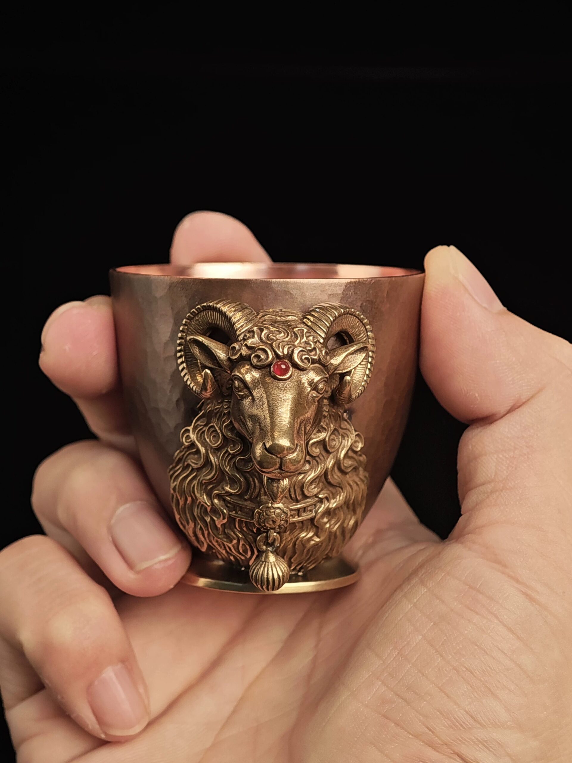 Hand-held scale view of Classic ram-head copper cup with pale-gold hanging bead against black background